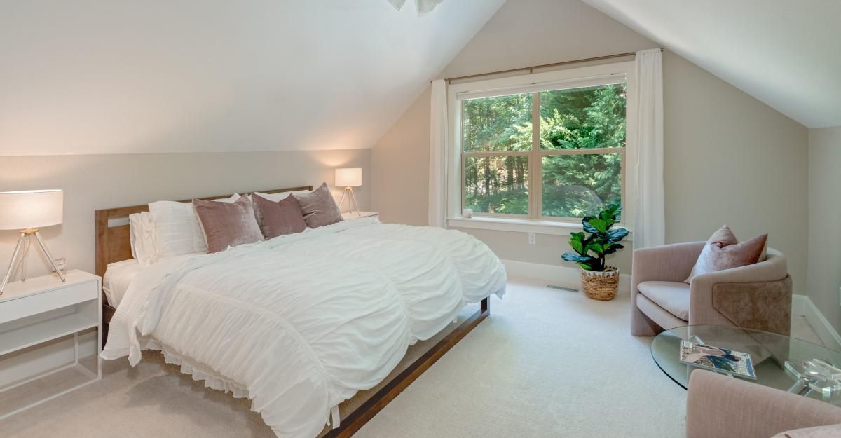 Bedroom with a large bed, a window with greenery, and a pink armchair. The walls are a soft gray, and the room is well-lit.