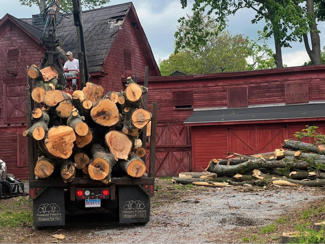 A truck filled with logs is parked in front of a red barn