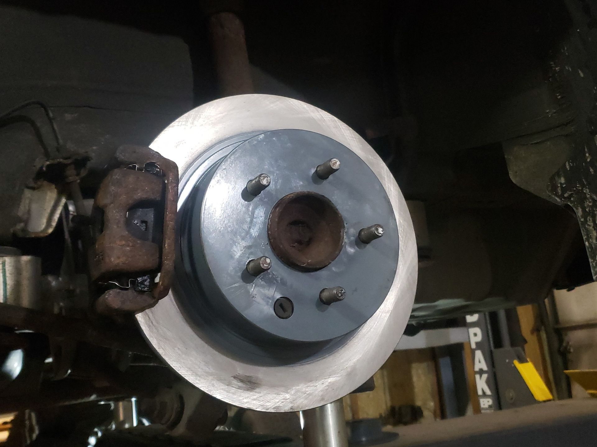 Car brake rotor and caliper assembly; grey rotor, rusty caliper. Underside view, repair shop.