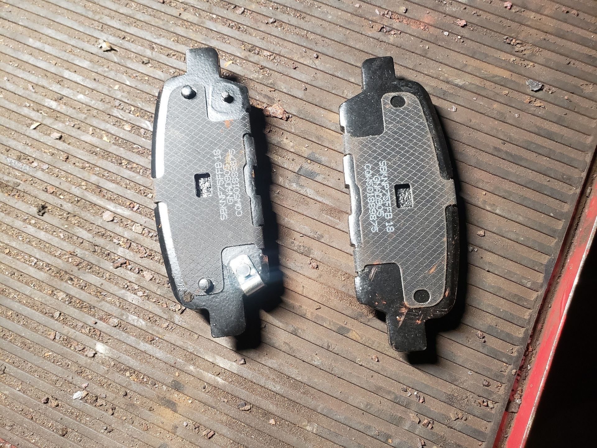 Two used brake pads, black and gray, lying on a metal surface with brown debris.