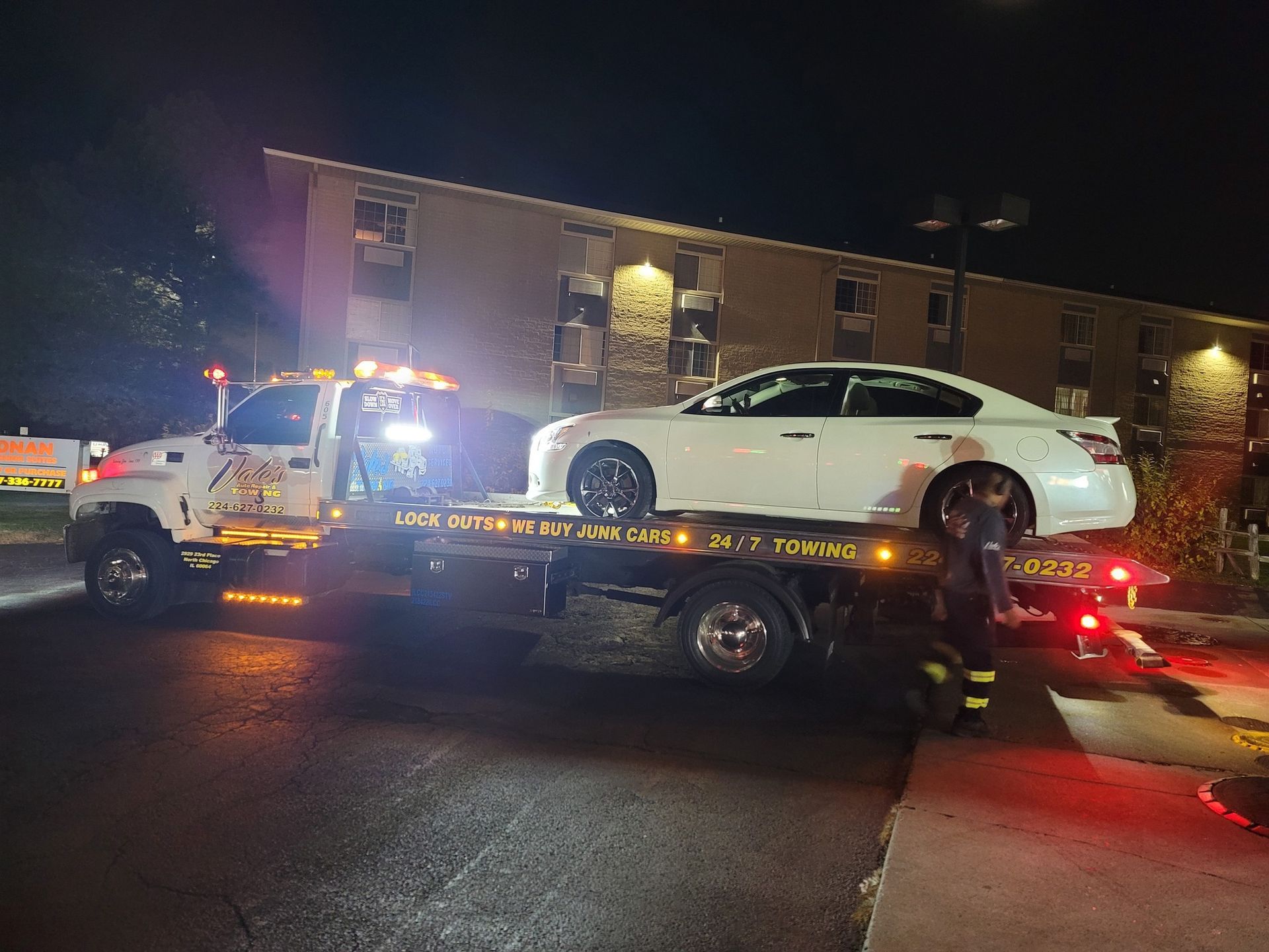 A white car being loaded onto a tow truck at night, in front of a building.