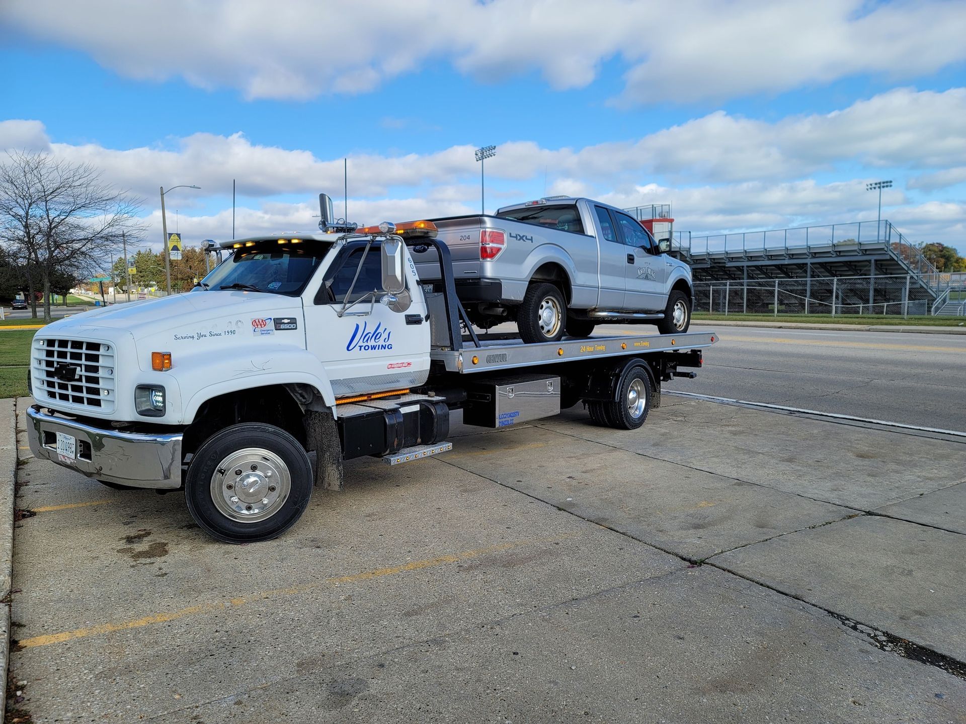 White tow truck carrying a silver pickup truck in a parking lot. Blue sky.
