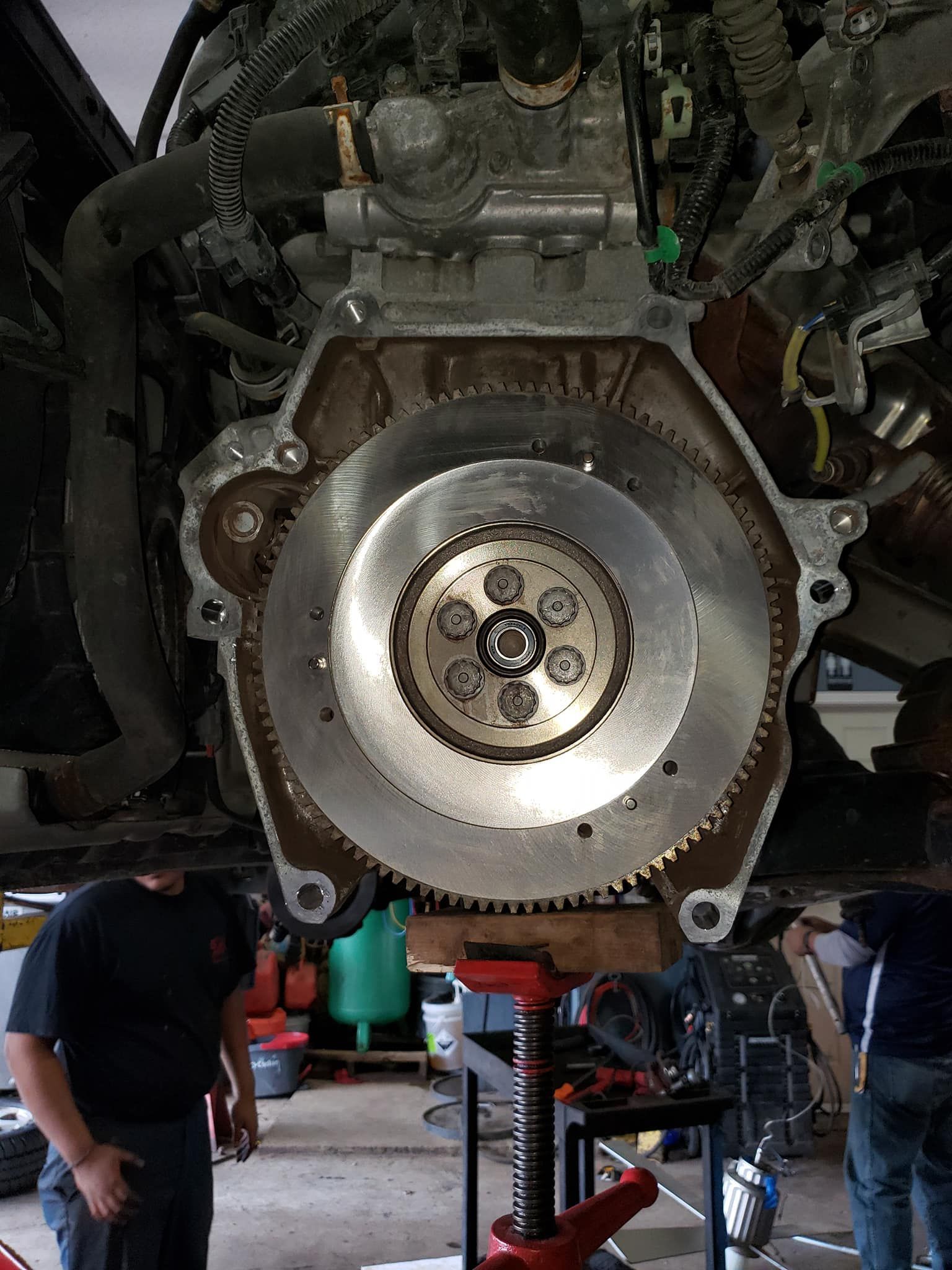 Engine with flywheel exposed, being worked on by two people in a garage.