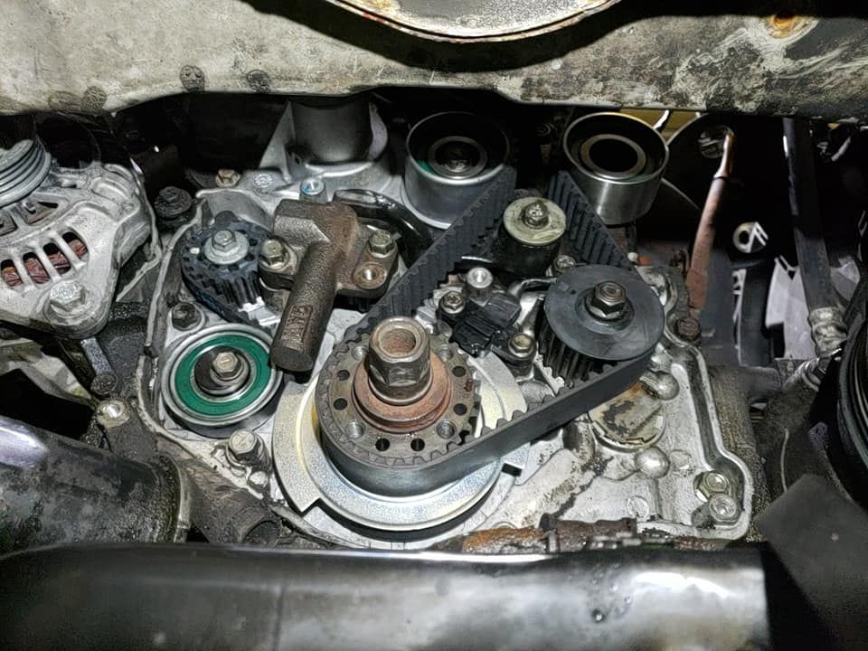Overhead view of an engine's exposed timing belt and pulleys. Gray metal, black belt, various pulleys, and bolts are visible.