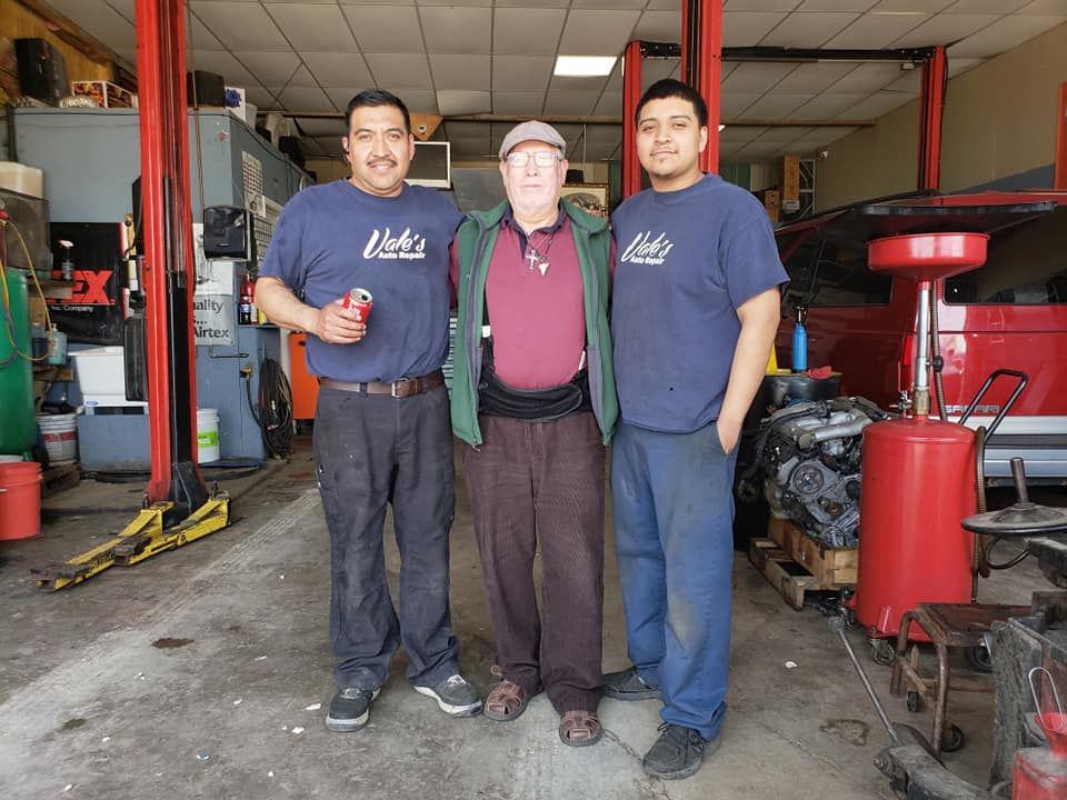 Three men posing in a car repair shop. Two in matching blue shirts with a logo, one in a green jacket.