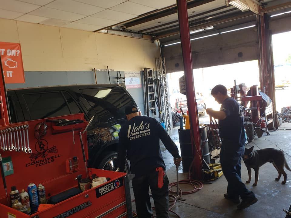 Two mechanics working on a car inside a repair shop, dog nearby. Tools are visible.