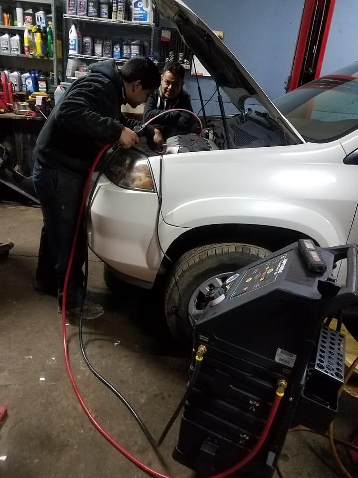 Two mechanics work on a white car with its hood open, using equipment in a garage.