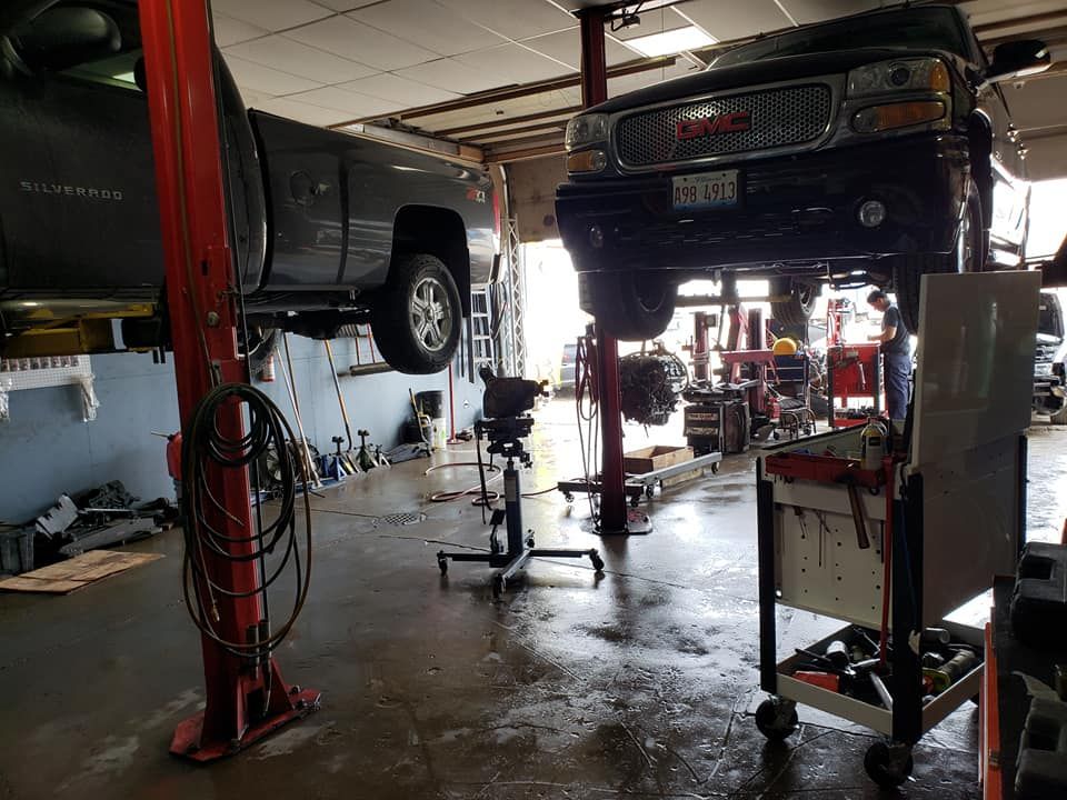 Two trucks on lifts in an auto repair shop. Tools, equipment, and a mechanic are visible.