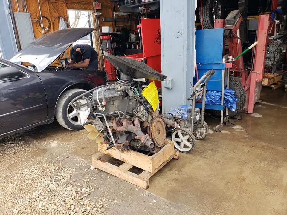 Car engine on a wooden pallet in a garage, with a mechanic working on a car in the background.