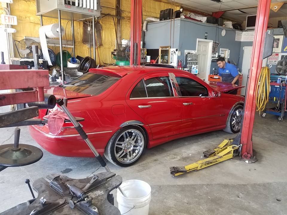 Red car being worked on in an auto repair shop. Mechanic in blue shirt nearby.