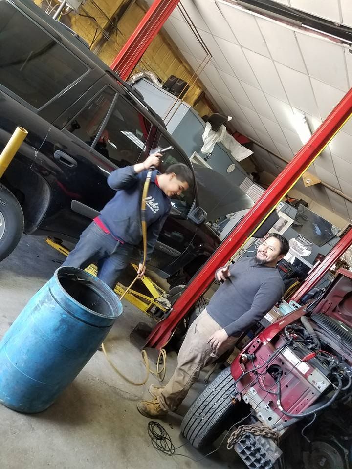 Two people working in a repair shop with cars on lifts, one pouring liquid, the other smiling.