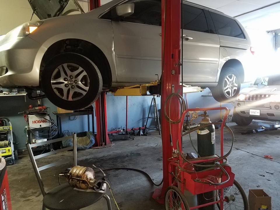 Silver minivan on a car lift in a garage. Mechanic work area with tools and equipment visible.