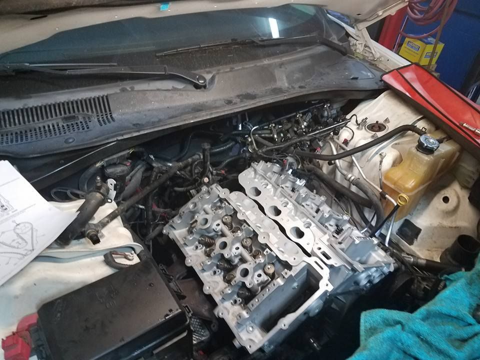 Car engine partially disassembled. Cylinder head removed, various parts scattered around engine bay.