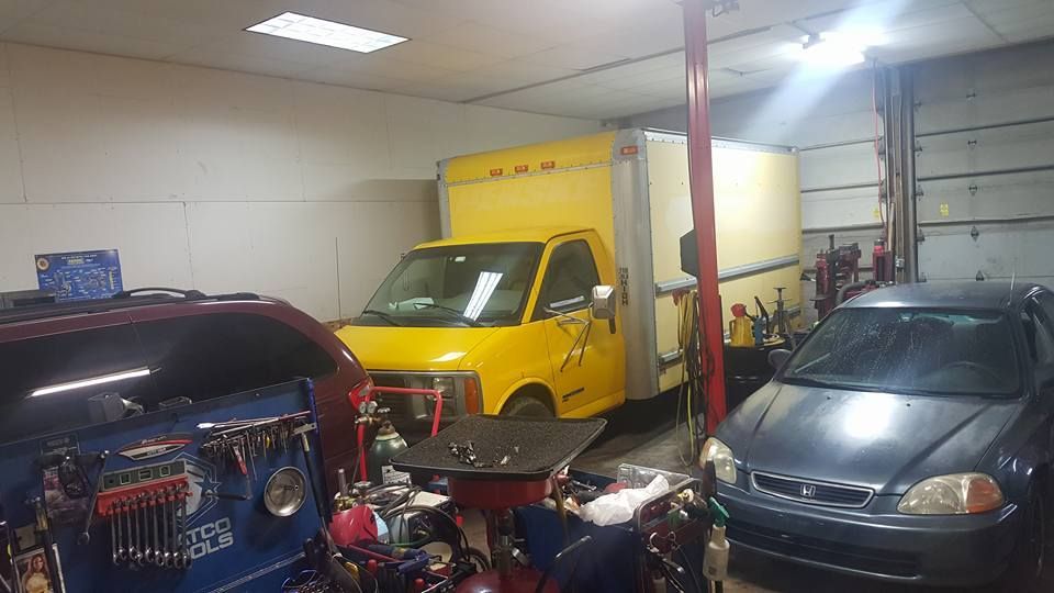 A cluttered garage with a yellow box truck, a red car, and a gray sedan. Tools and equipment are scattered about.