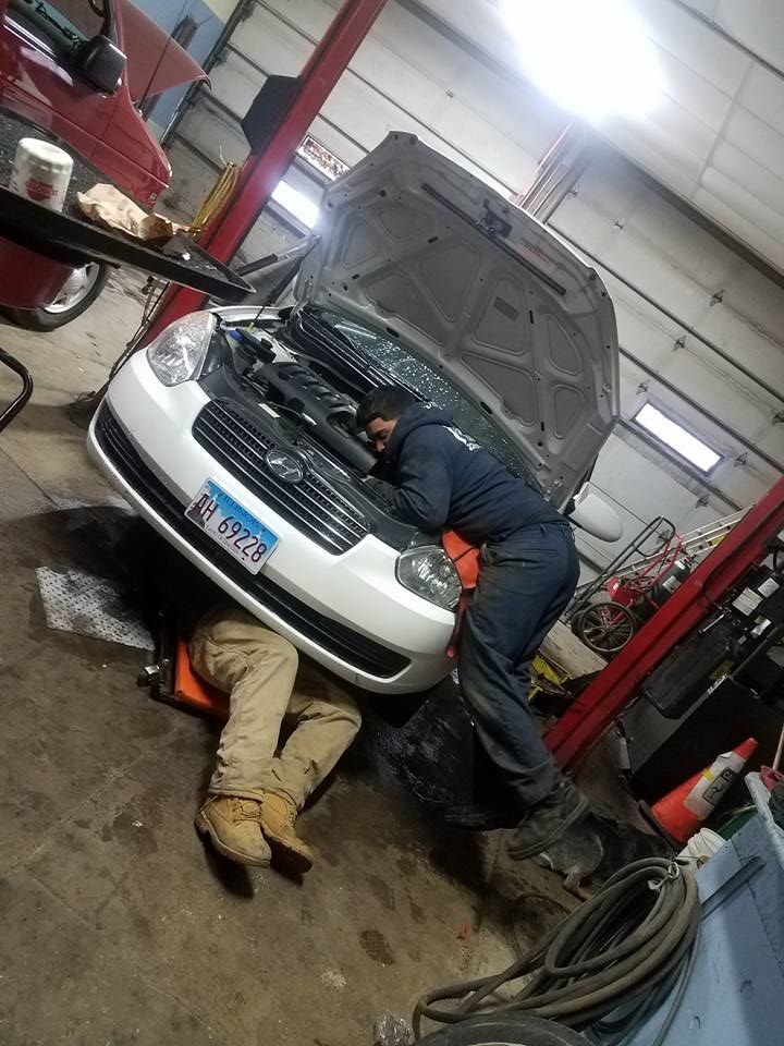 Two mechanics working on a white car in a garage; one under, one with hood open.