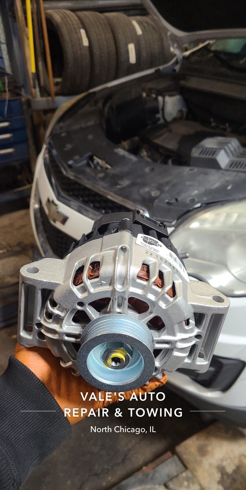 A person holding a car alternator, near an open car hood in a repair shop.