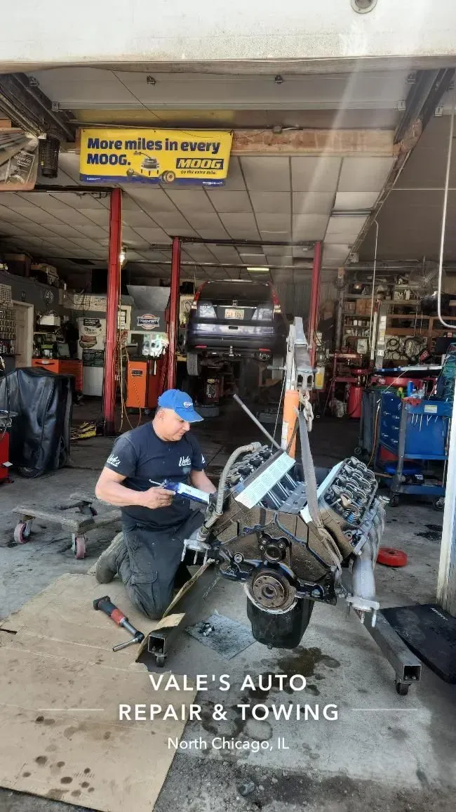 Mechanic working on an engine in a repair shop; gray concrete floor, signs, tools visible.