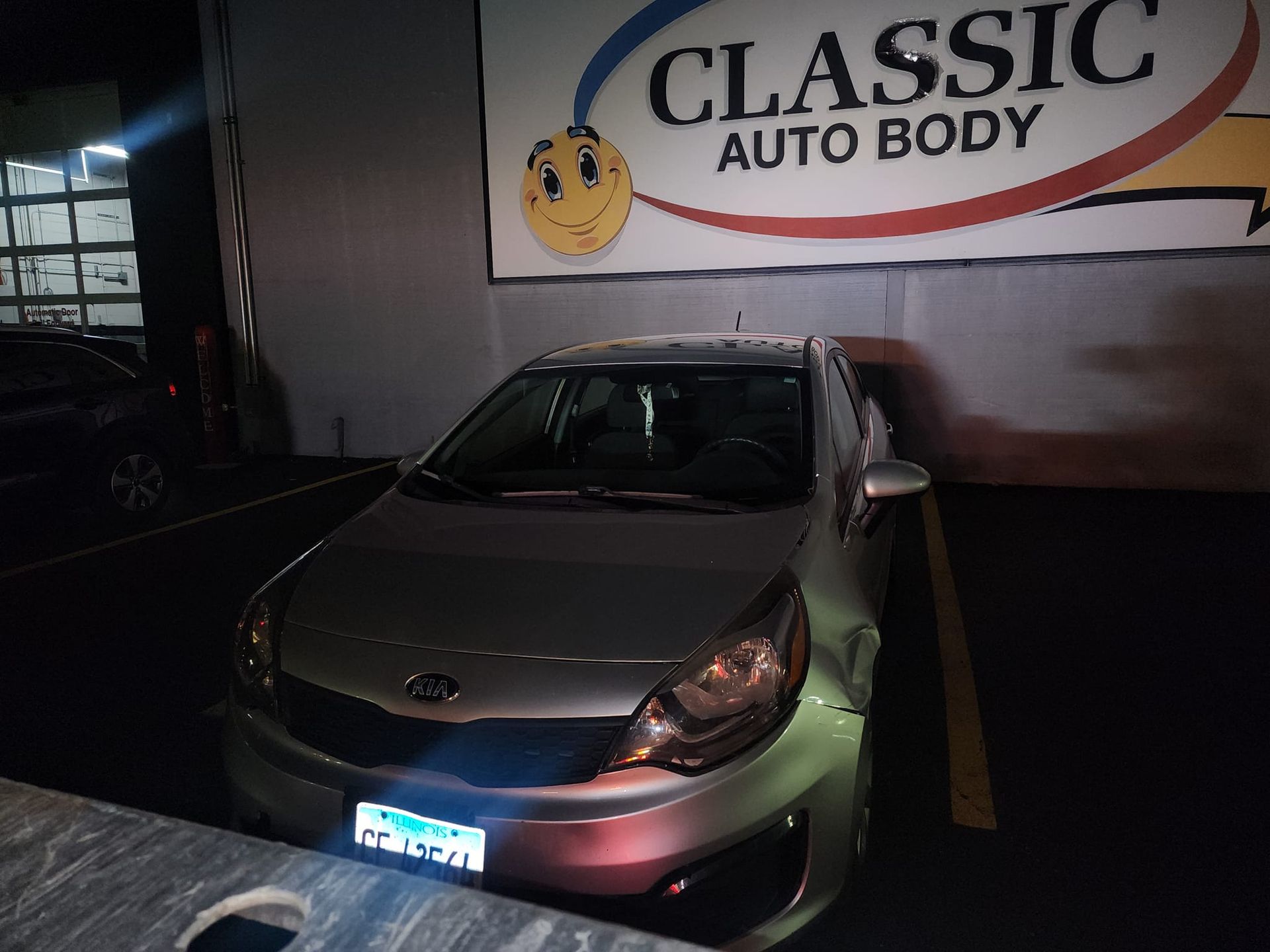 Silver Kia Rio parked in front of Classic Auto Body sign at night.