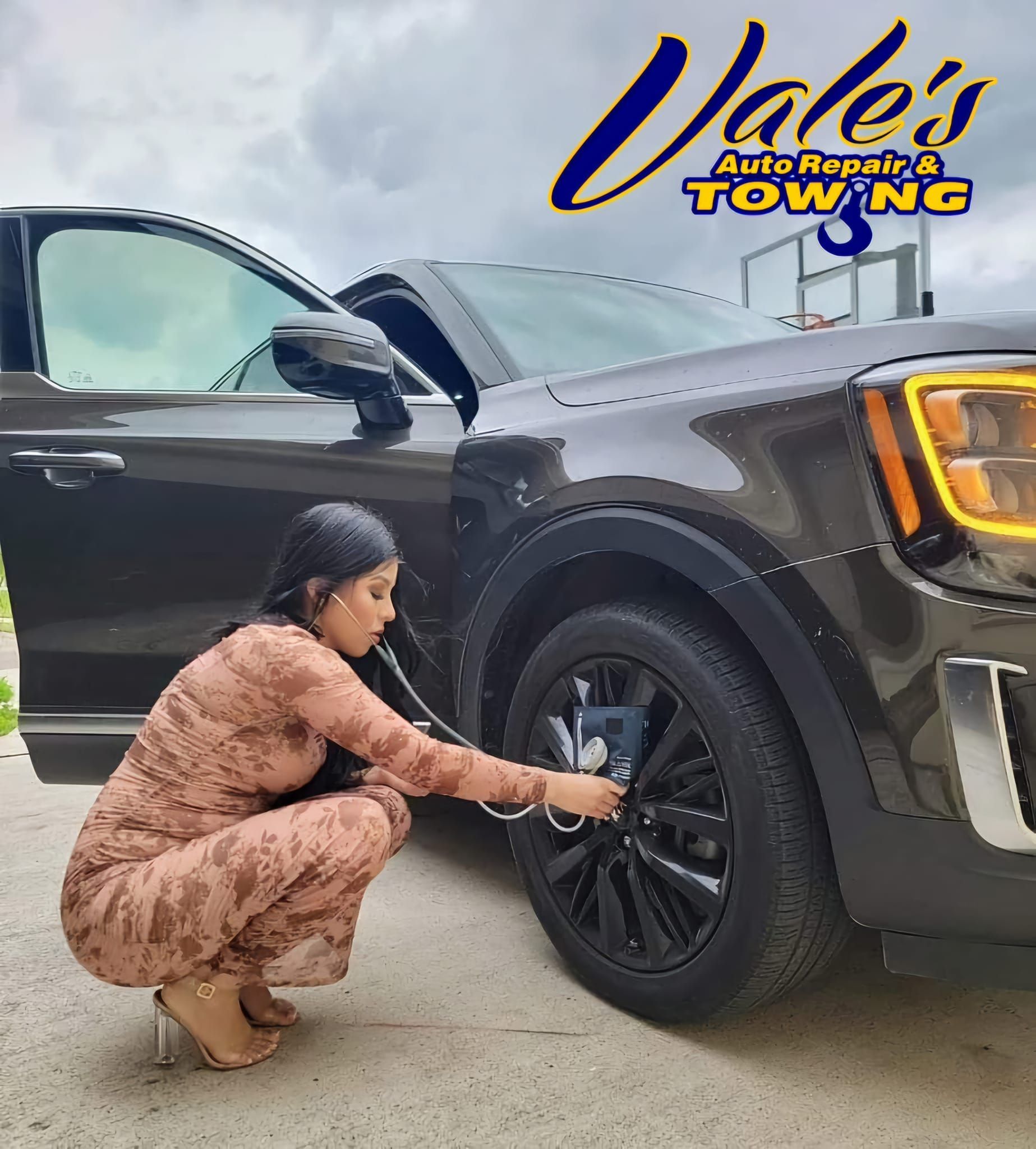 Woman kneeling by a dark SUV's tire, likely checking it. Vale's Auto Repair & Towing logo in the background.