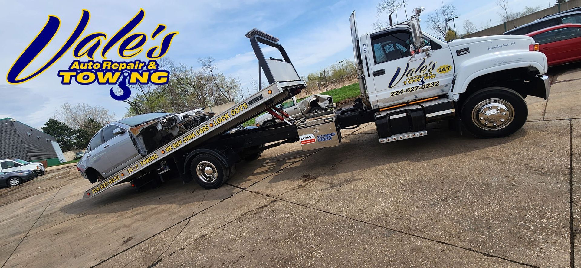 A white tow truck with a car on its flatbed is parked on a paved surface. The tow truck has 