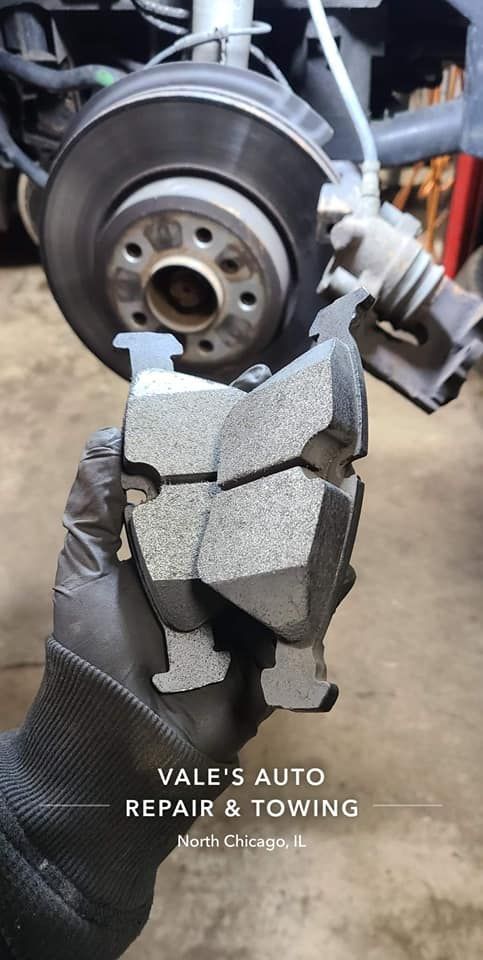 A hand in a black glove holds used brake pads in front of a car's brake rotor.