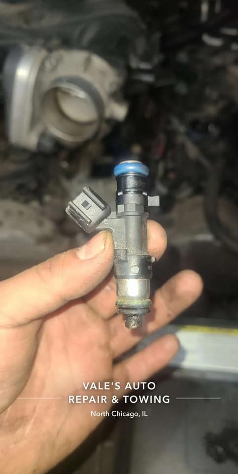 A hand holds a fuel injector with a blue cap. The setting is a car engine bay, with other engine parts in the background.