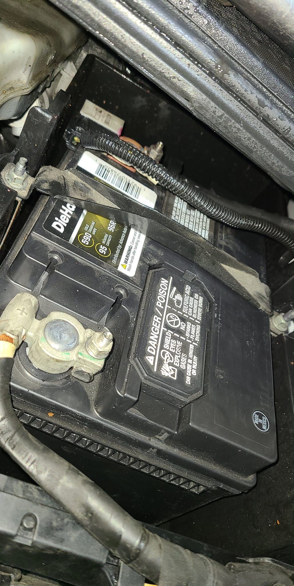 Car battery in engine compartment, surrounded by wires and components.
