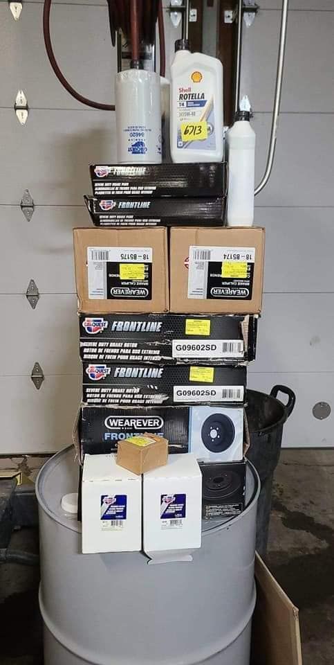 A stack of car parts on a metal drum includes oil, filters, and brake pads in a garage setting.