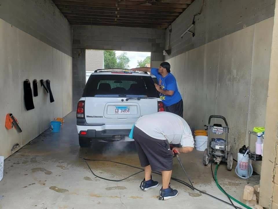 Two people washing a white SUV in a car wash bay. One rinses the back, the other cleans the top.