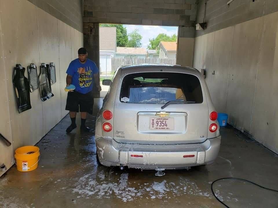 Man washing a silver car at a car wash. Foamy water, yellow bucket, open air, sunny day.