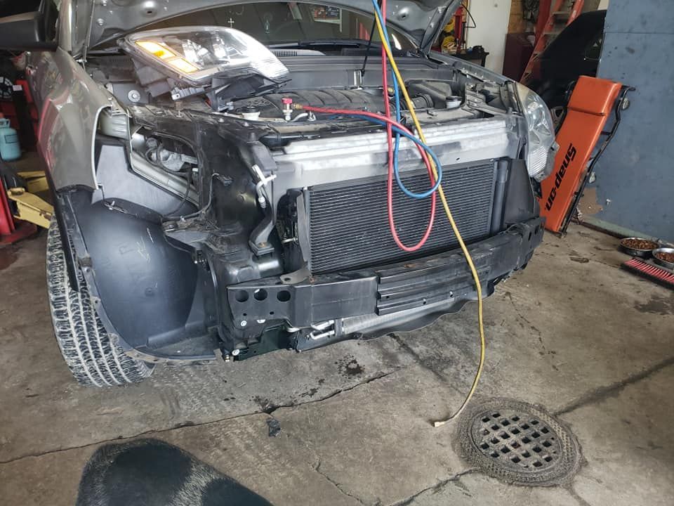 Car with front-end damage, being repaired with hoses connected. Hood open in a garage setting.