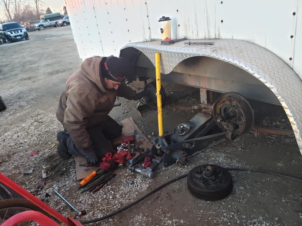 Person working on a trailer's axle outdoors. Tools and parts are scattered around, with the wheel removed.