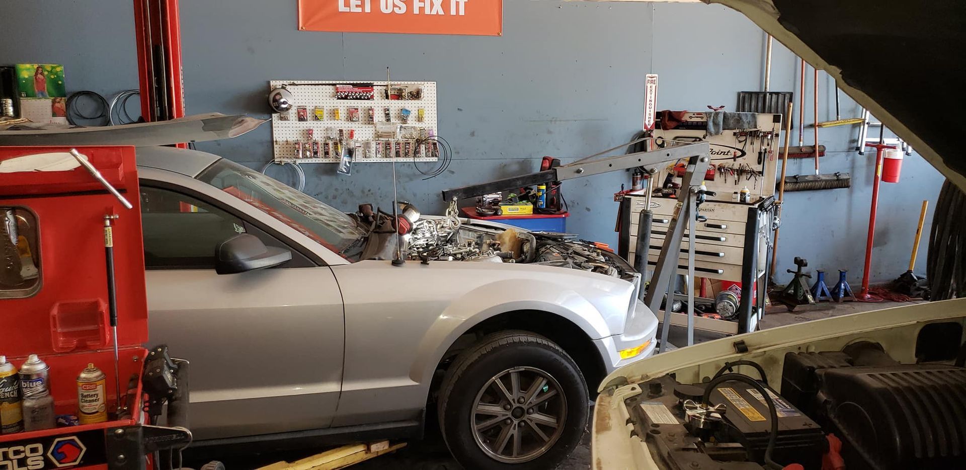 Silver car in a repair shop with its hood open; another car's hood is visible on the right. Tools and equipment are scattered around.