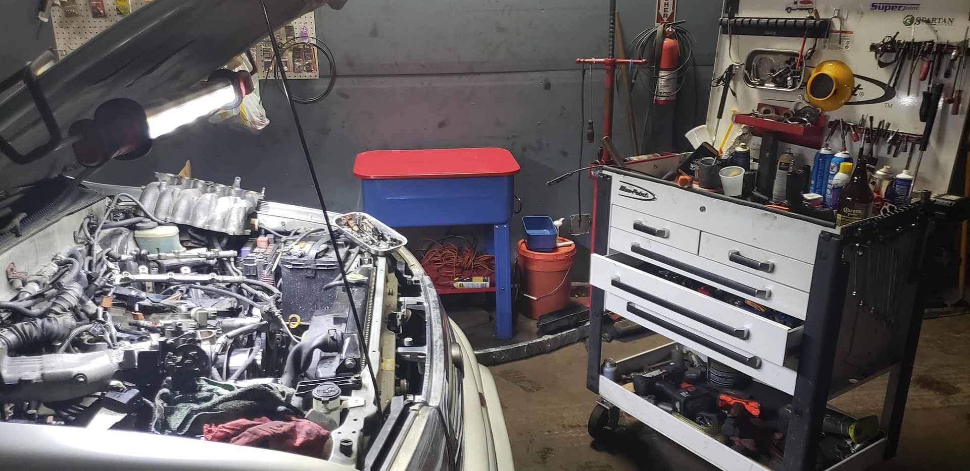 Car engine bay open in a cluttered mechanic shop with a tool cart.