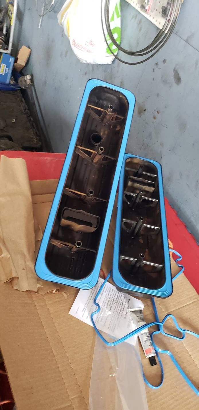 Two blue-trimmed valve covers with debris and a new blue gasket on a cardboard surface.