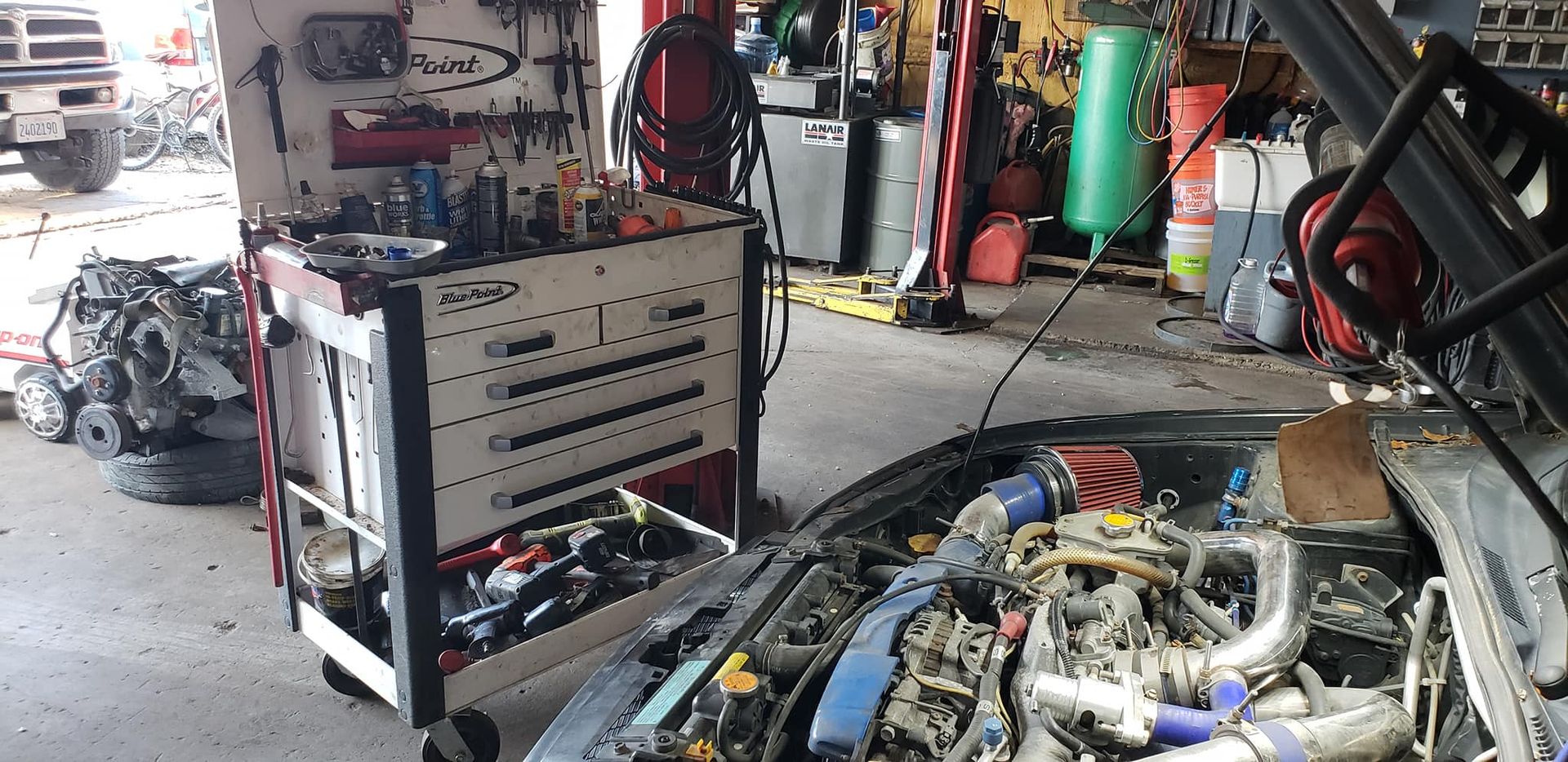 Car engine under repair in a garage, tools and equipment visible, including a toolbox and air intake.