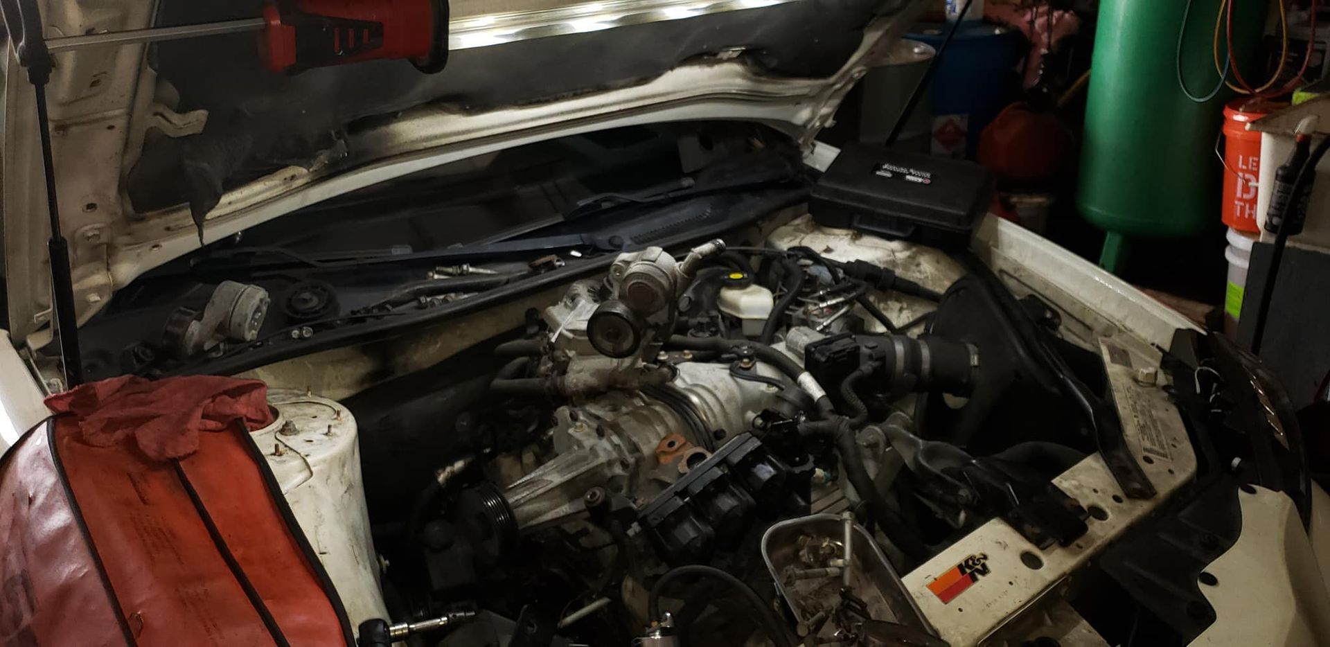 Engine of a white car with the hood open, likely undergoing repair in a garage setting.