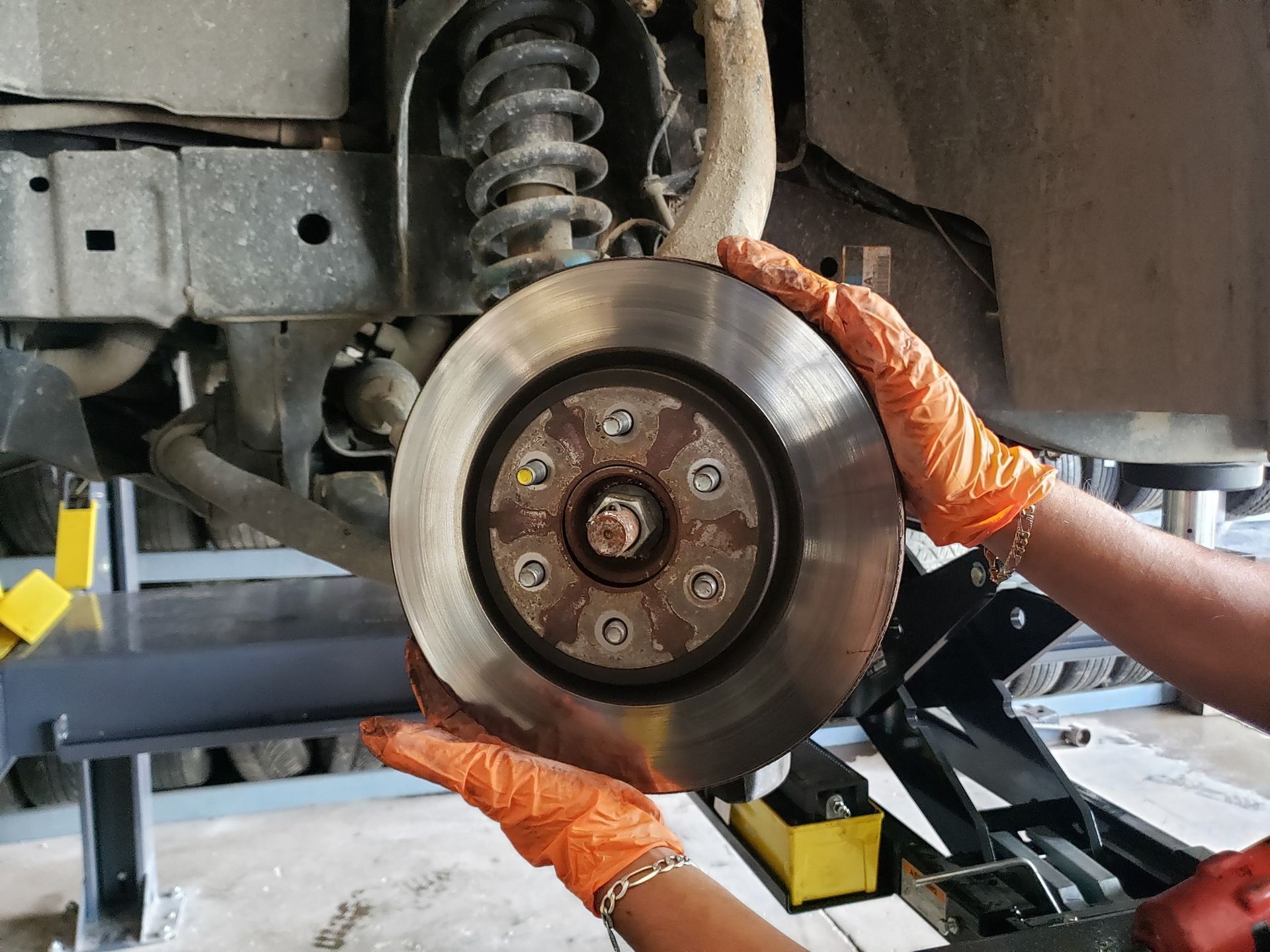 Hands wearing orange gloves holding a brake rotor next to a car's suspension.