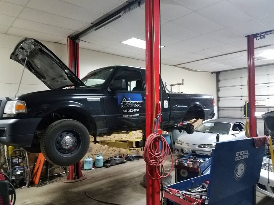 Black truck on a lift in a repair shop with its hood open, tools and other vehicles visible.