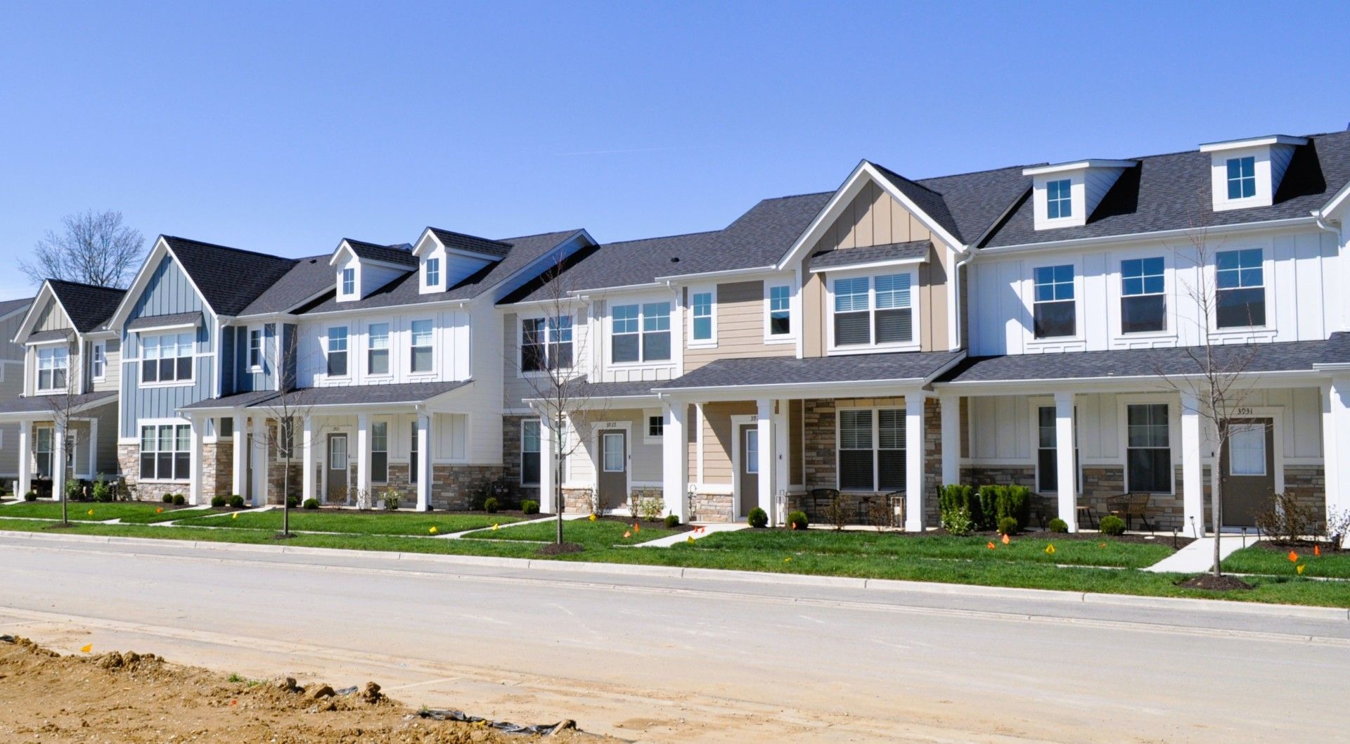 A row of newly built, multi-story townhomes with white siding, dark roofs, and stone accents along a paved road.