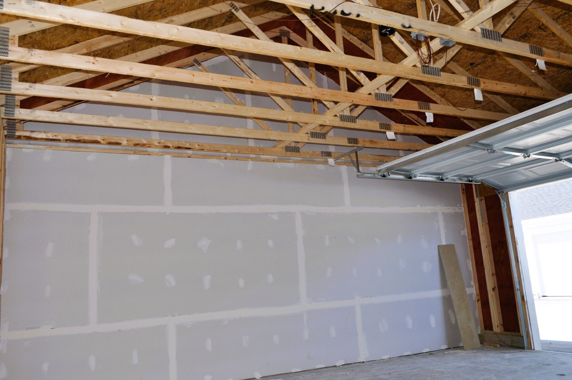 Interior of a garage under construction featuring newly installed gray drywall sheets on a wall below exposed wood trusses.