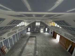 Interior of a large room under construction with drywall on walls and ceiling, windows, and bare concrete floor.
