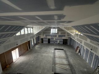 Interior construction of a large room with drywall on walls and ceiling. Windows and open doorways visible.