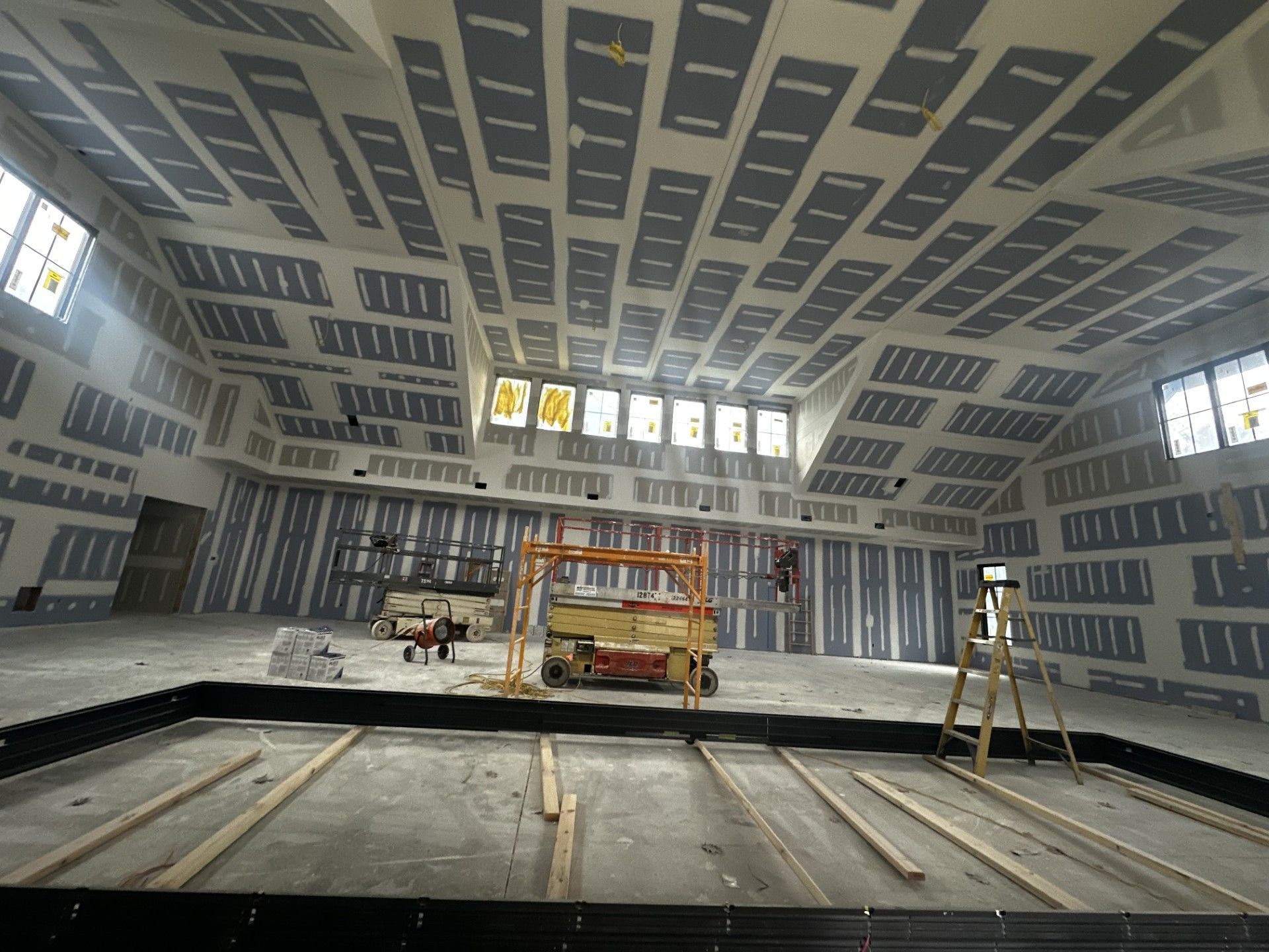 Interior construction site with drywall installation on walls and ceiling; a lift and ladder are present.