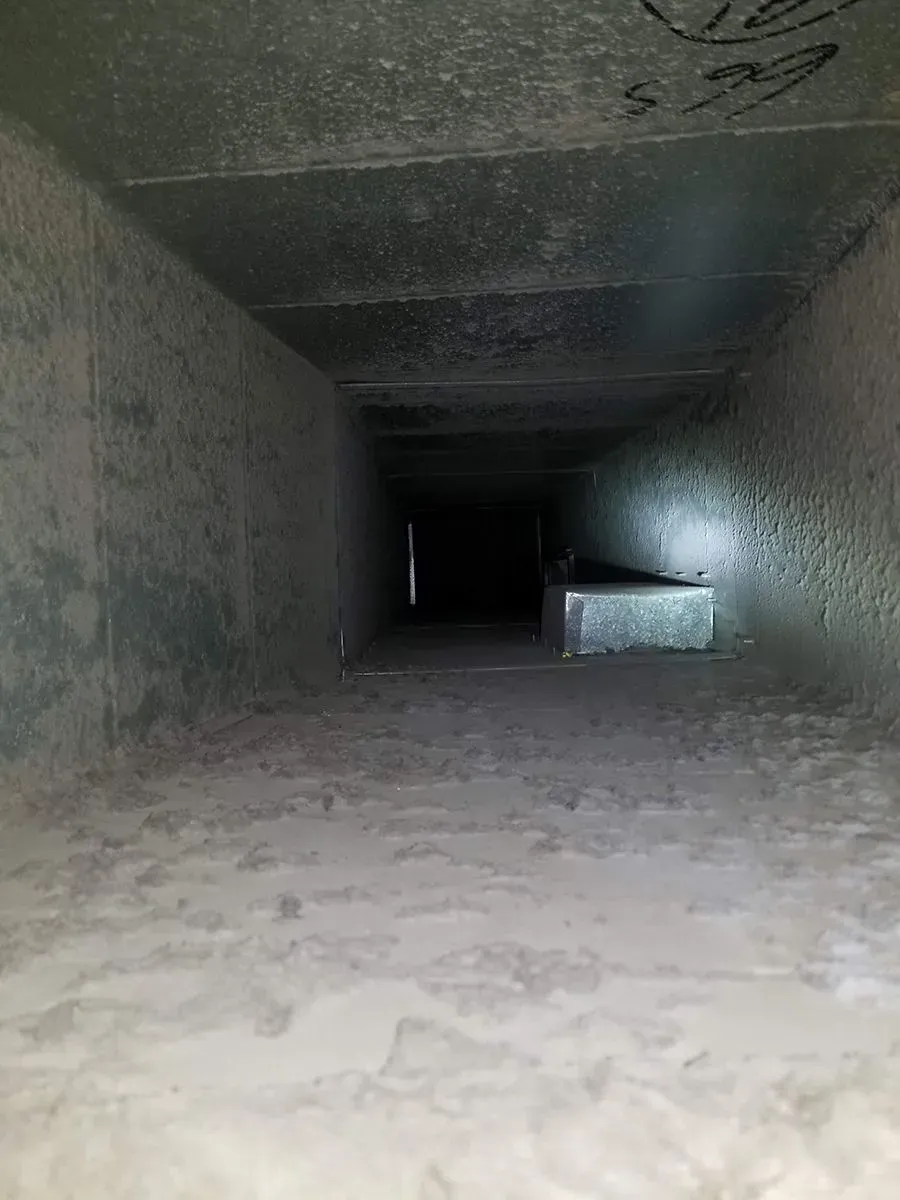 Inside of a rectangular, dusty duct. A light shines on a metal component in the dark distance.