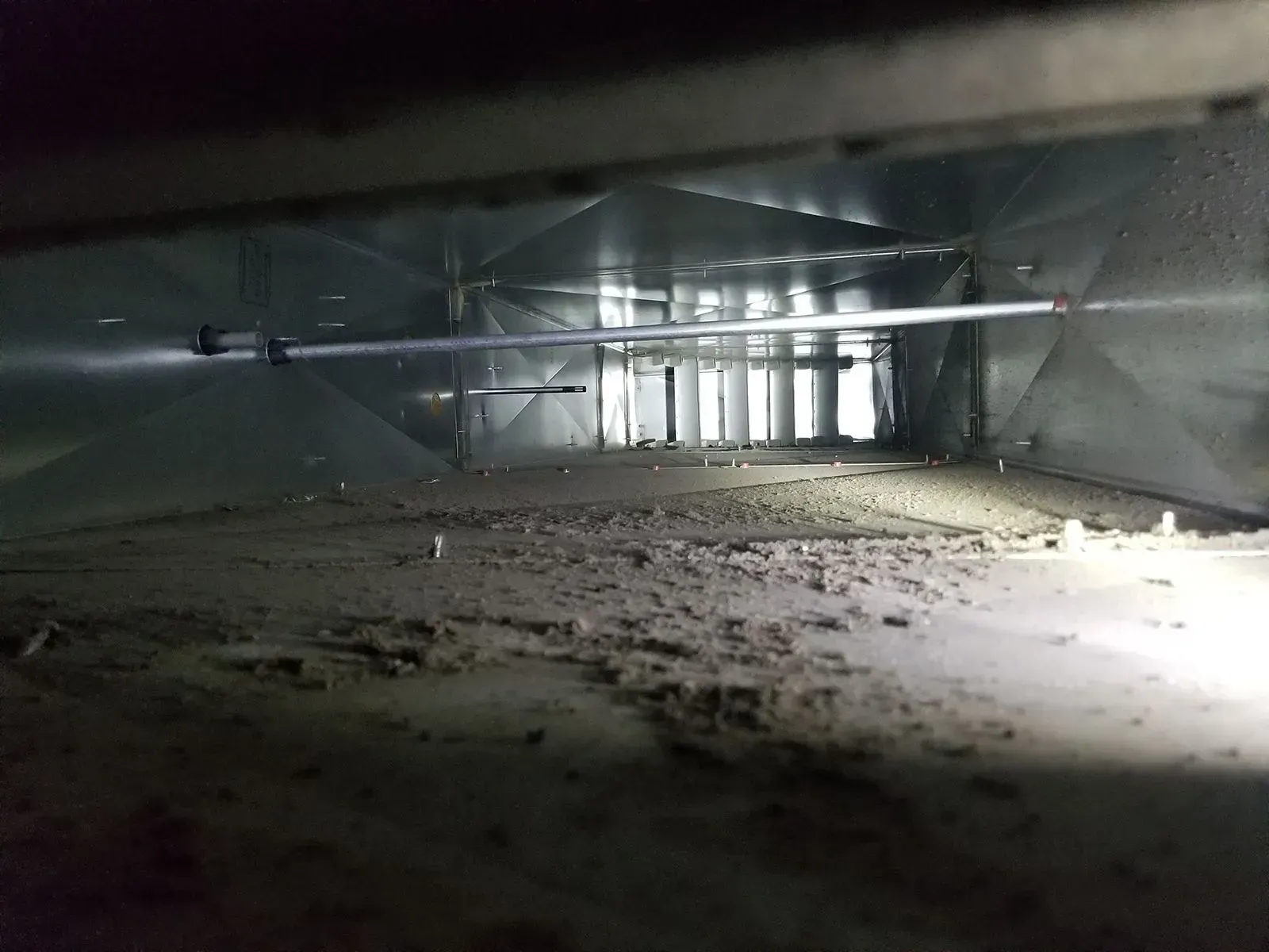 Inside view of a dusty HVAC duct. Silver metal interior, with visible debris on the bottom. Light source at the end.