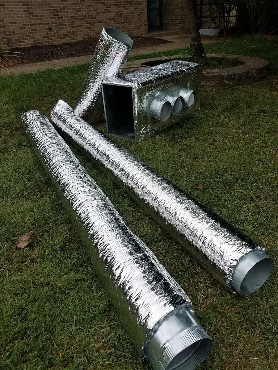 Silver HVAC ductwork and a rectangular air handler box rest on grass outdoors.