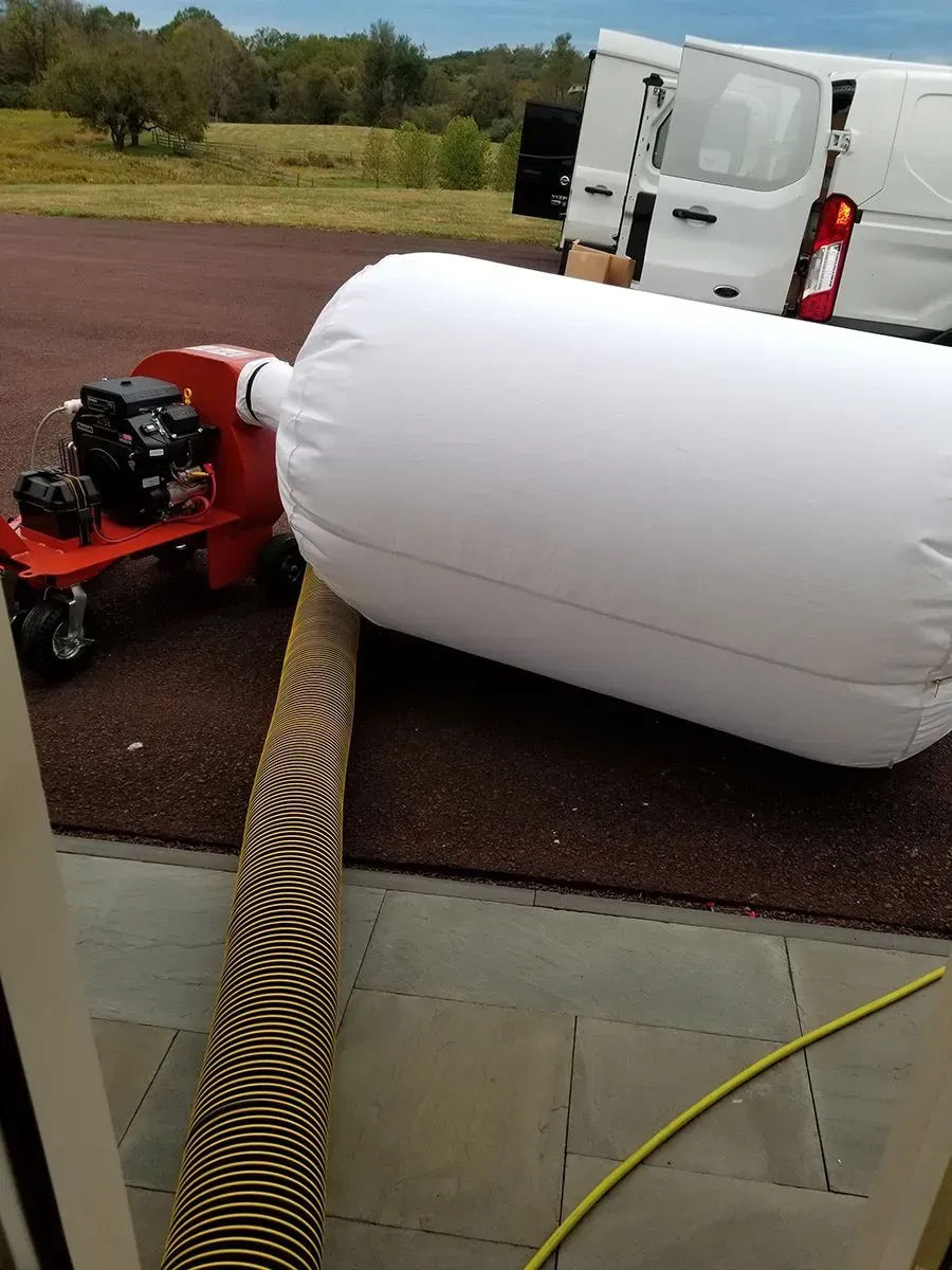 White inflatable connected to a blower on wheels, next to a van.  Brown ground and gray patio.