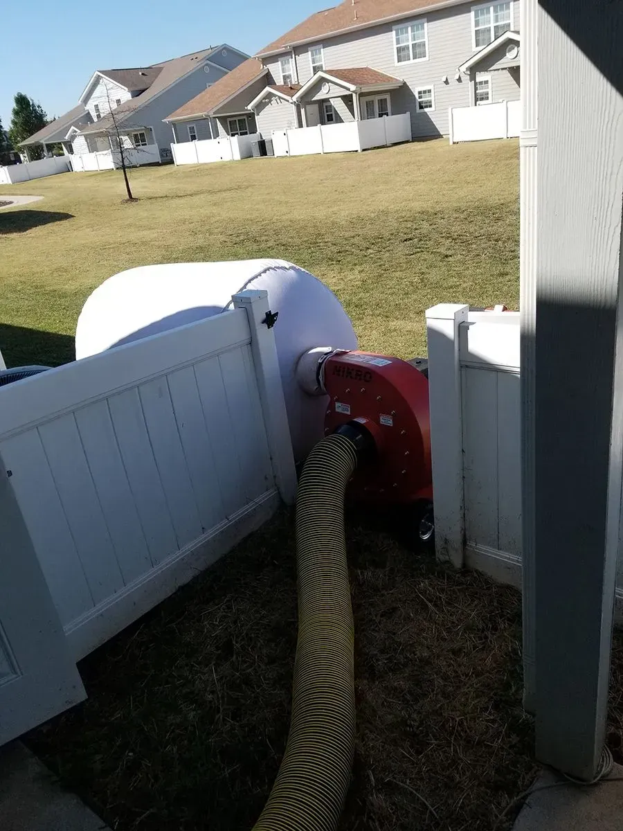 White fence with hose connected to red device, grass, and houses in background.