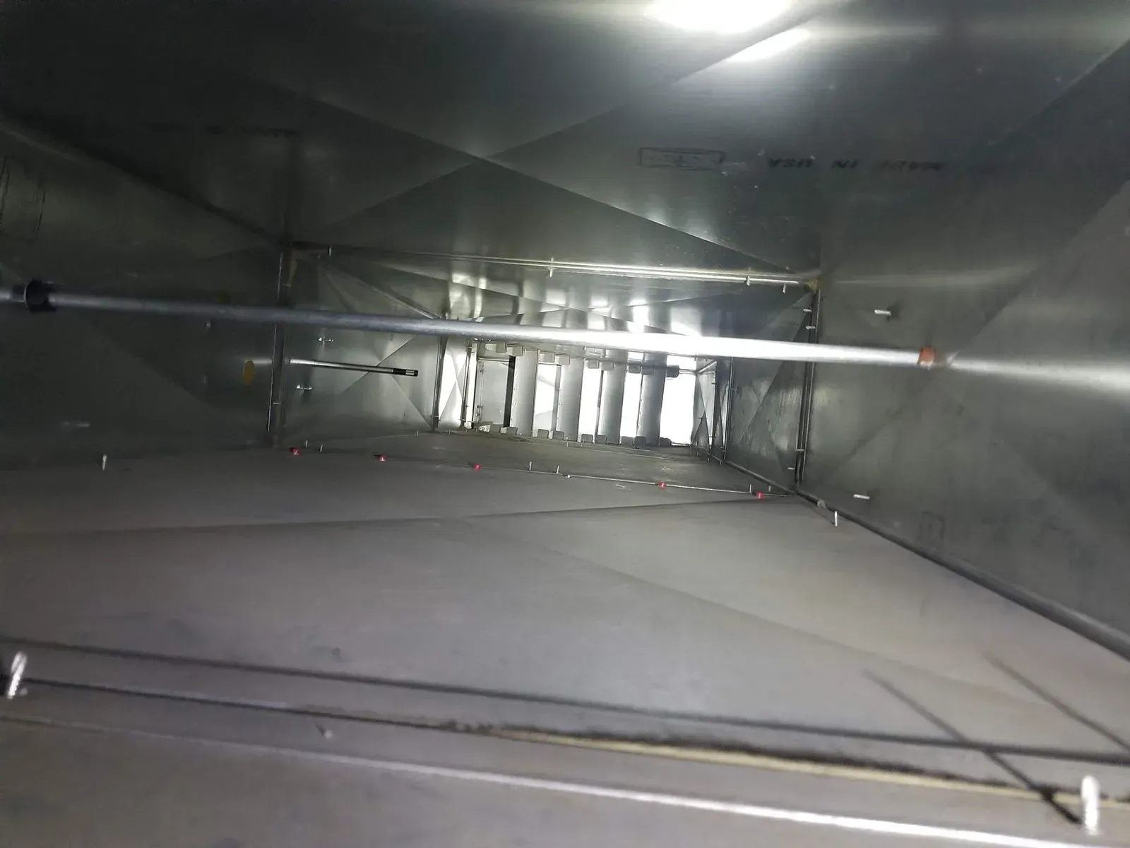 Inside a shiny, rectangular HVAC duct, a metal pipe spans the width, with open slots visible towards the end.